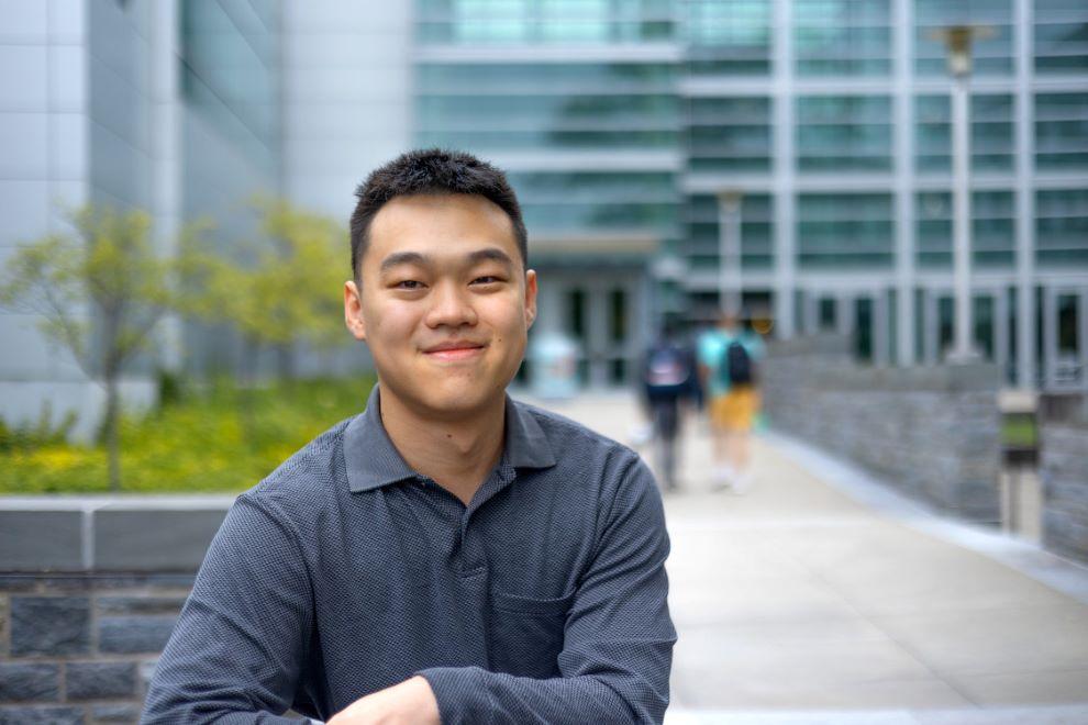 Student in blue shirt smiling in front of a walkway and building